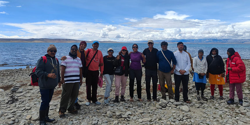 A group of tourists posing on the rocky shore of Lake Mansarovar in Tibet with scenic mountains and cloudy sky in the background.