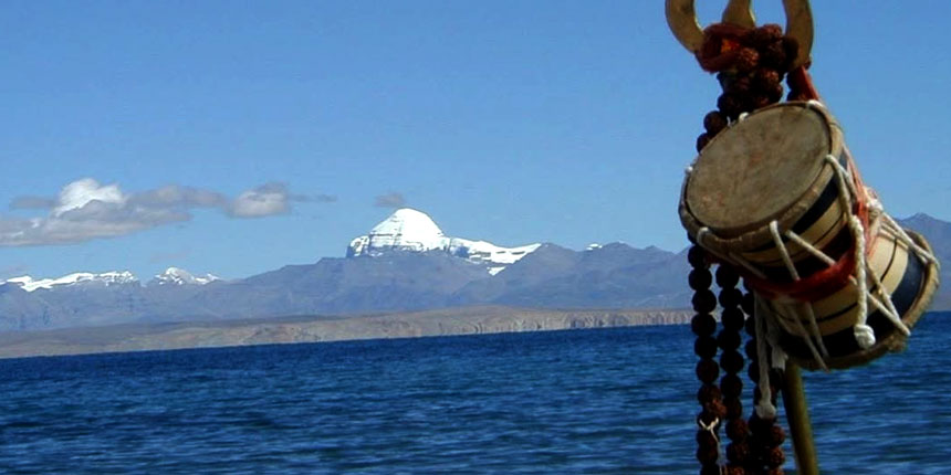 A close-up of a damaru, a two-headed drum associated with the Hindu deity Shiva, hangs in the foreground with a string of prayer beads. In the background, the vast blue waters of Lake Manasarovar lead to the snow-covered peak of Mount Kailash under a bright sky.