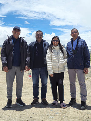 Four standing together outdoors under a bright sky during a trip kailash yatra, smiling at the camera.