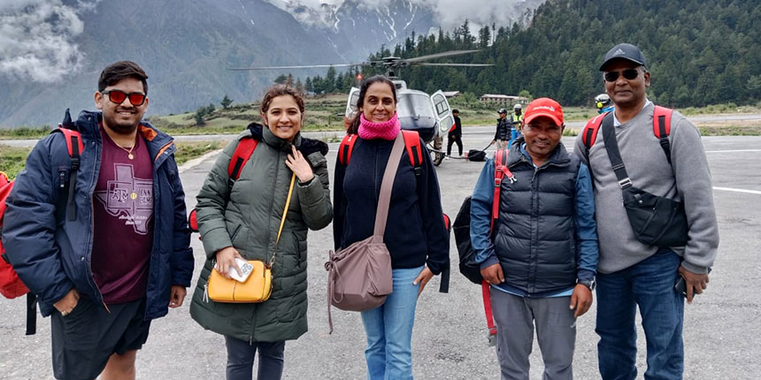 A group of pilgrims standing on a helipad with backpacks, smiling before boarding a helicopter for the Kailash Manasarovar Yatra, with mountains in the background.