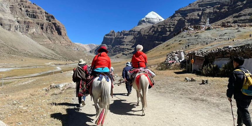 Pilgrims on horseback riding along a mountain trail during the sacred Mount Kailash Parikrama, with the snow-capped peak of Mount Kailash in the background.