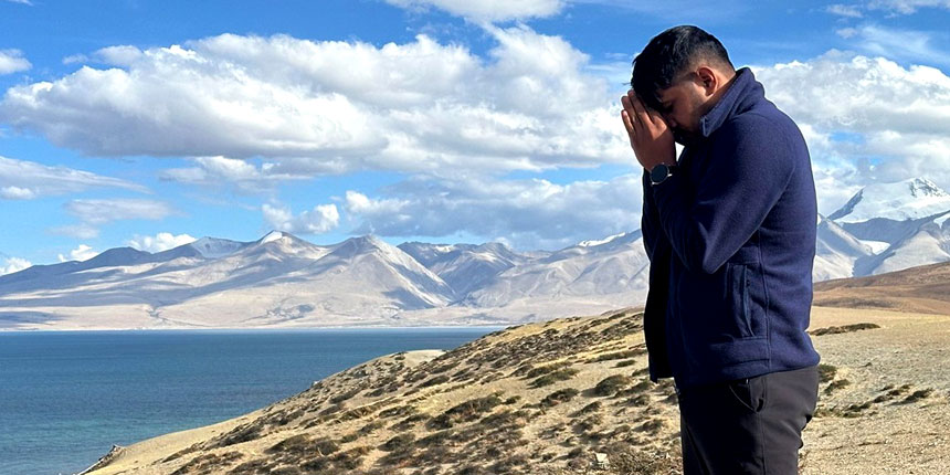 A man praying with folded hands near a serene lake and snow-capped Himalayan mountains in the background