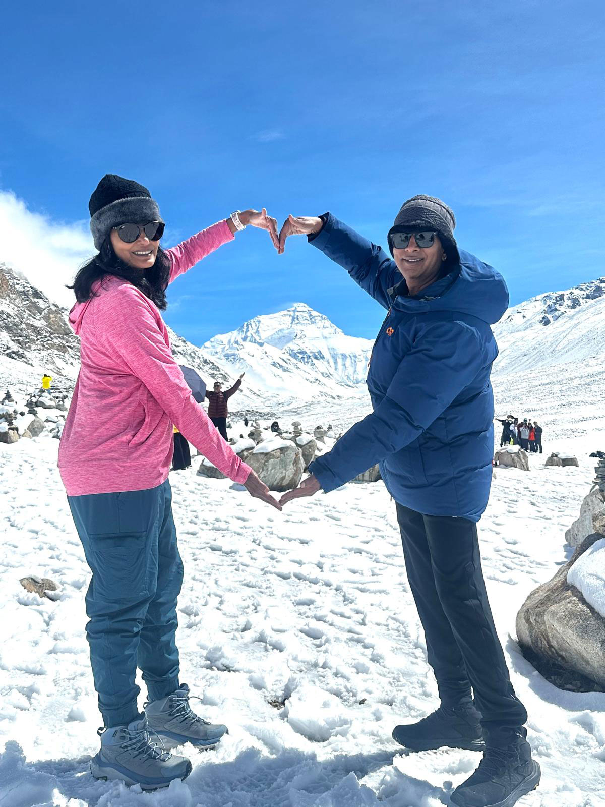 A smiling couple standing on snow-covered terrain, forming a heart shape with their hands against the backdrop of Mount Kailash under a clear blue sky.