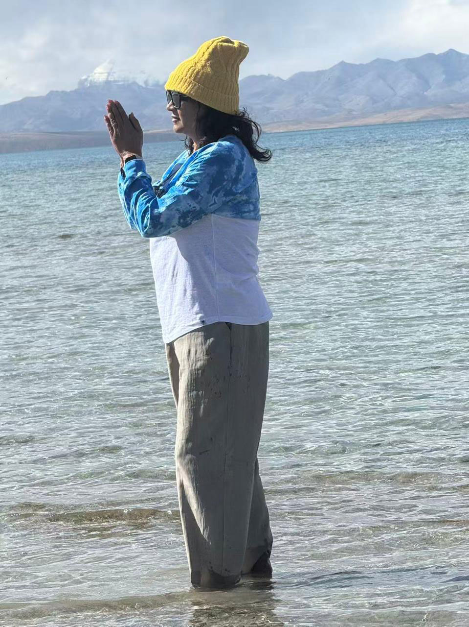 A woman standing in the waters of Lake Manasarovar with folded hands in prayer, wearing a yellow cap and blue-white top, with Mount Kailash visible in the distance.