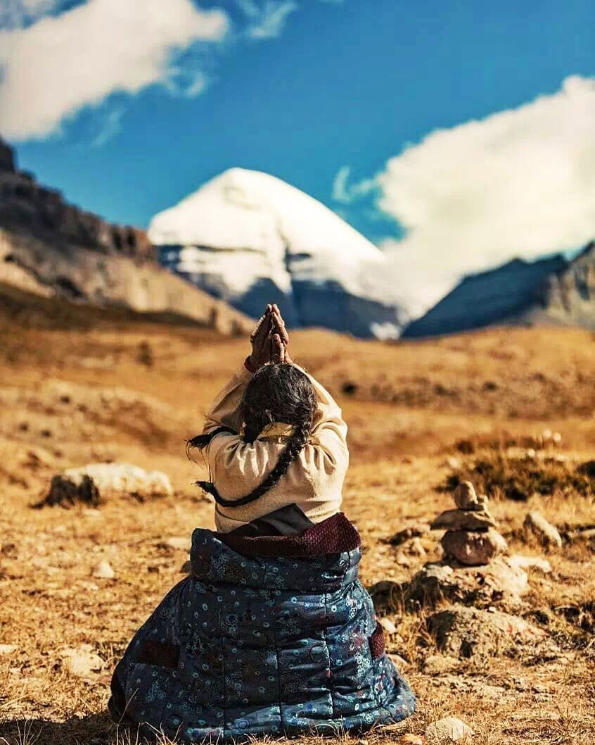 Tibetan woman with braided hair kneeling and praying with folded hands facing Mount Kailash, under a clear Himalayan sky