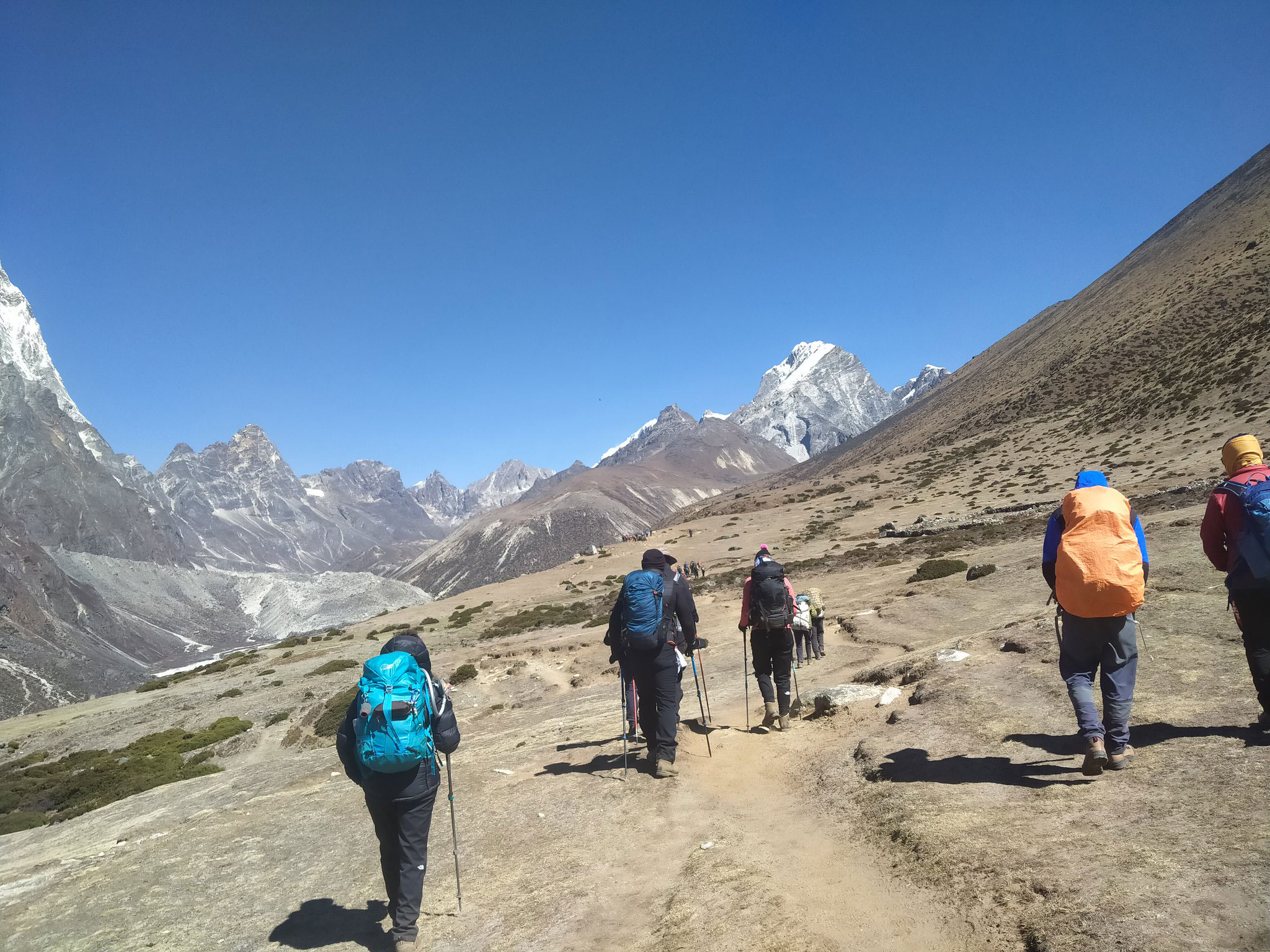 Group of hikers trekking towards Everest Base Camp with snow-capped Himalayan peaks in the background under a clear blue sky.