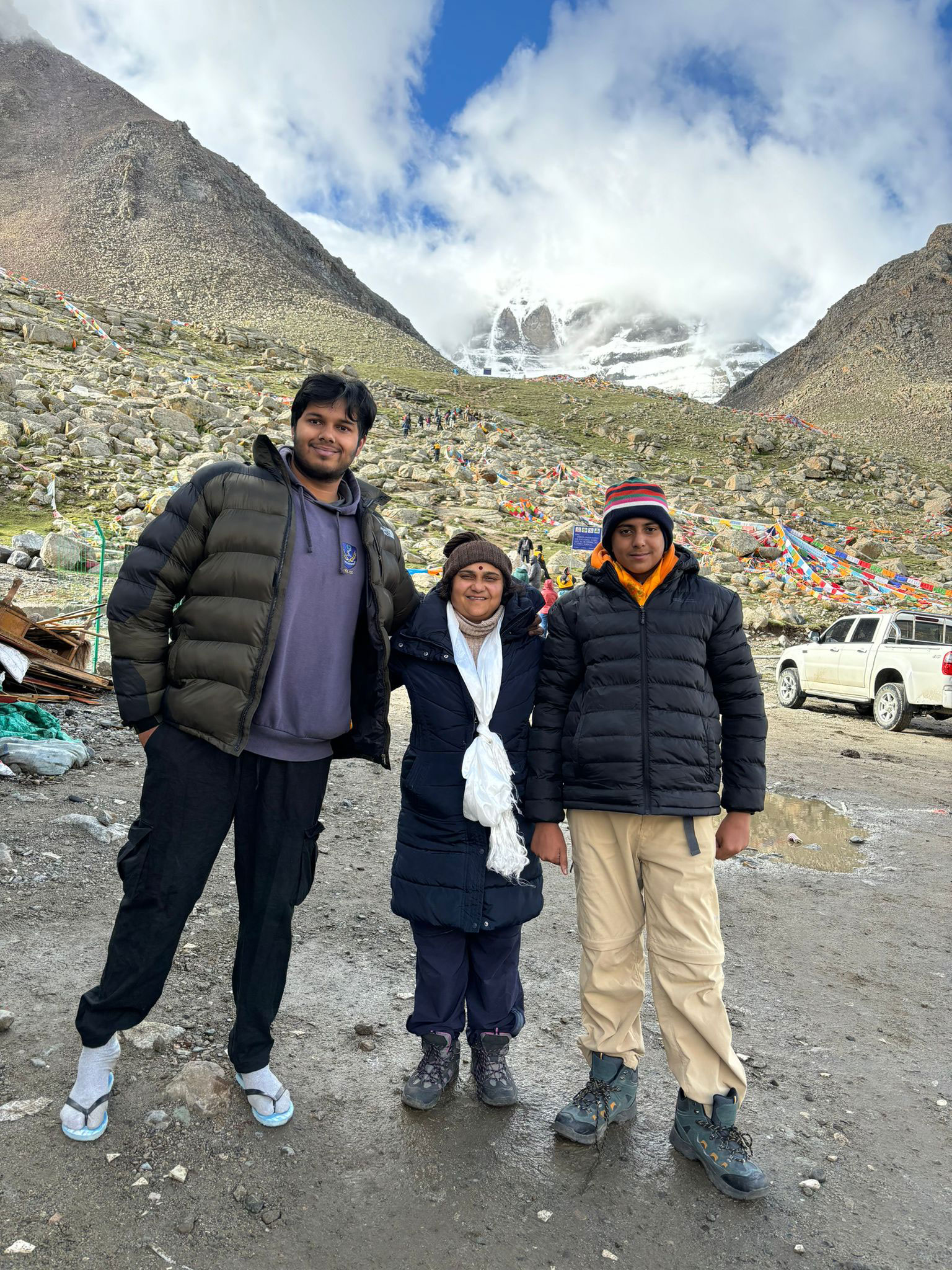 A family standing together and smiling in front of Mount Kailash, with colorful Tibetan prayer flags and snow-covered peaks in the background.