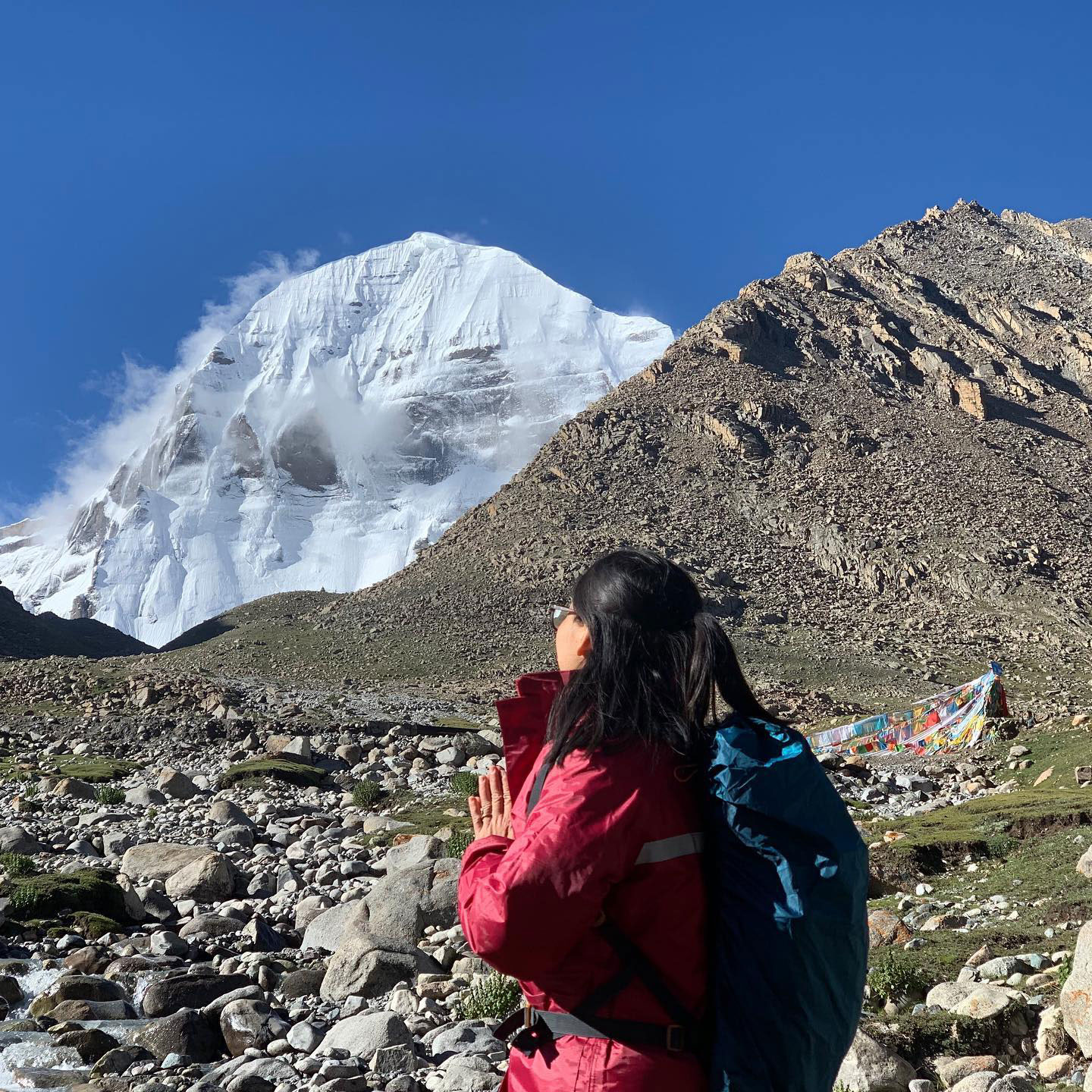 Woman trekker praying with folded hands facing Mount Kailash, with Tibetan prayer flags and rocky Himalayan terrain in the background
