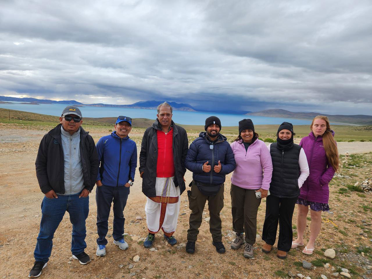 A group of travelers and pilgrims posing for a photo in front of the turquoise waters of Lake Manasarovar, with cloudy skies and mountain ranges in the background.
