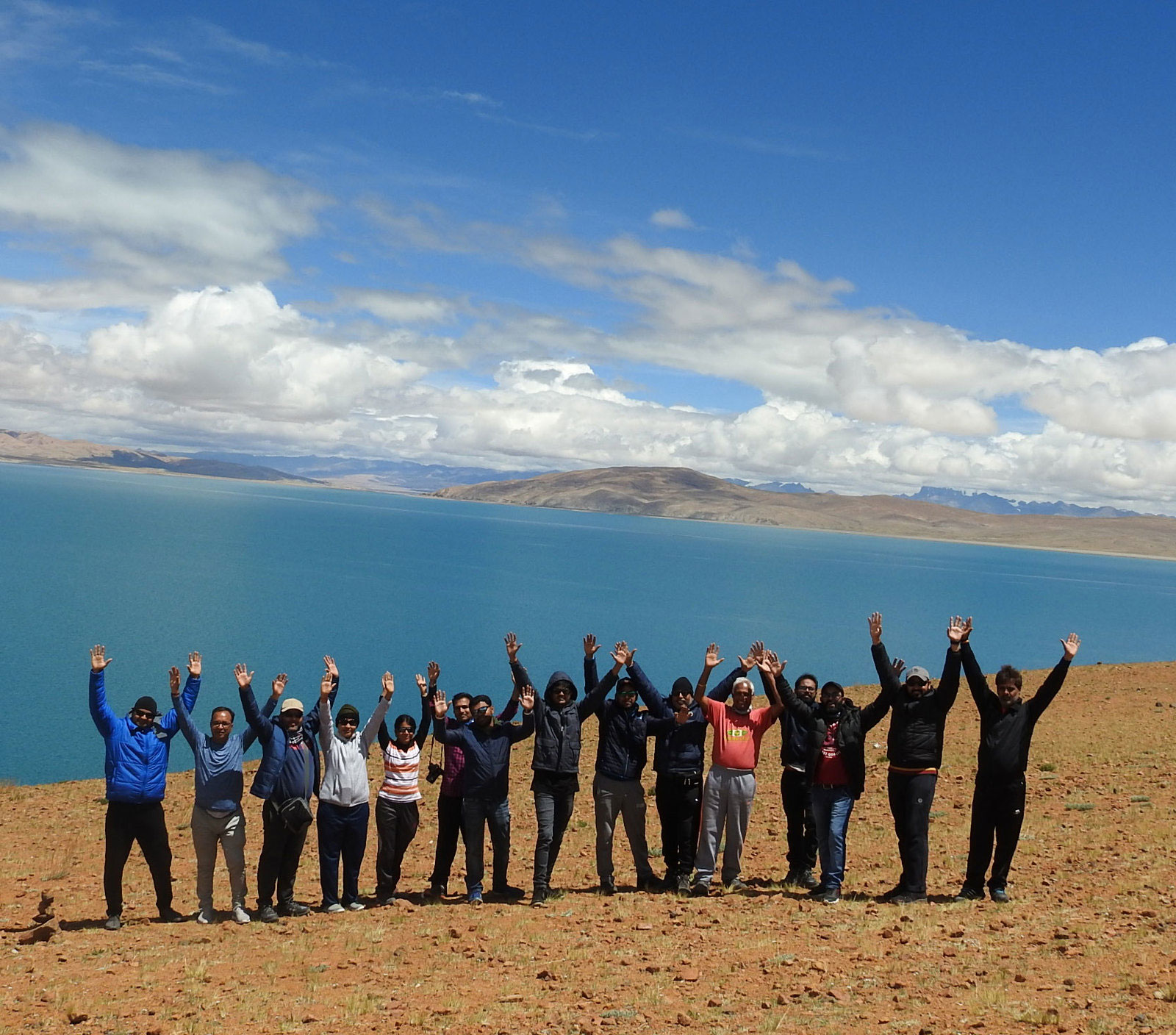 Group of pilgrims raising hands with joy at the scenic Mansarovar Lake during Kailash Mansarovar Yatra in Tibet, with clear blue sky and mountains in background
