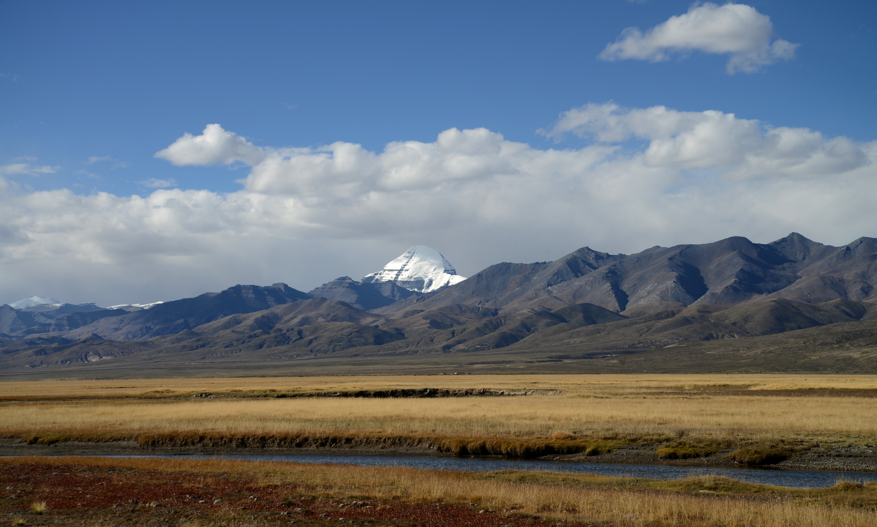 Mount Kailash seen from a distance across golden Tibetan grasslands with a small river in the foreground and rugged Himalayan mountains under a blue sky with scattered clouds.