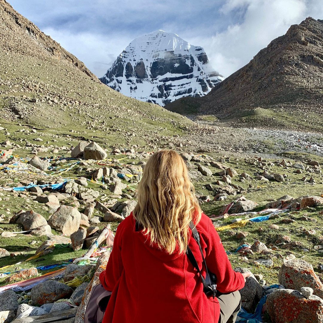 A devotee meditating in front of Mount Kailash during Kailash Mansarovar Yatra, surrounded by mountains, rocks, and Tibetan prayer flags.