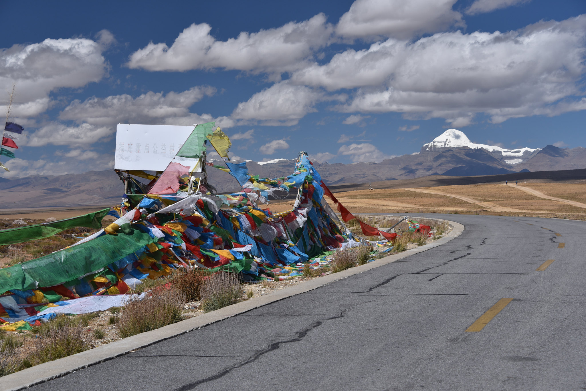 A winding road leading to Mount Kailash with colorful Tibetan prayer flags fluttering in the wind under a blue sky with clouds and a vast Himalayan landscape.