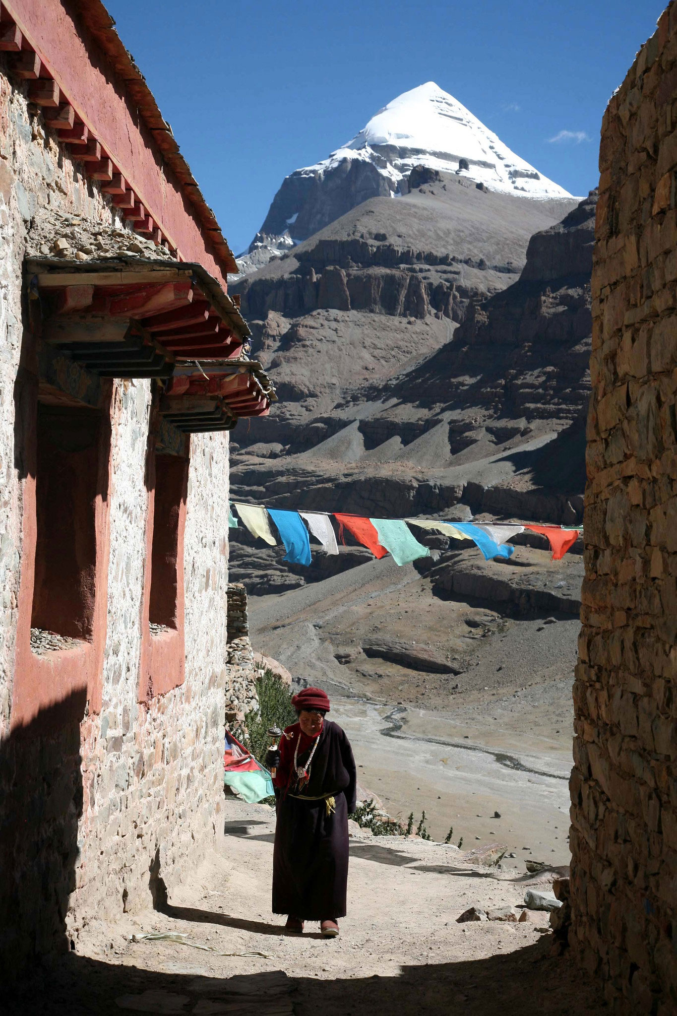 A Tibetan monk walking near traditional stone houses with colorful prayer flags, and the snow-capped Mount Kailash in the background.