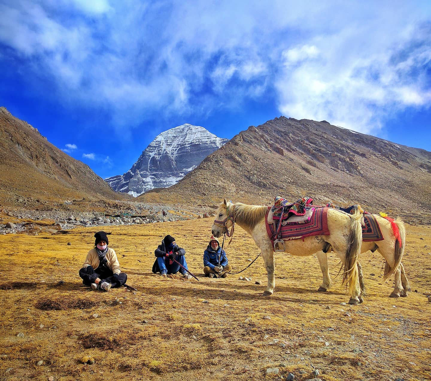 Three travelers resting with a decorated horse in the Himalayan plateau near Mount Kailash, with snow-covered peaks and dramatic skies in the background.