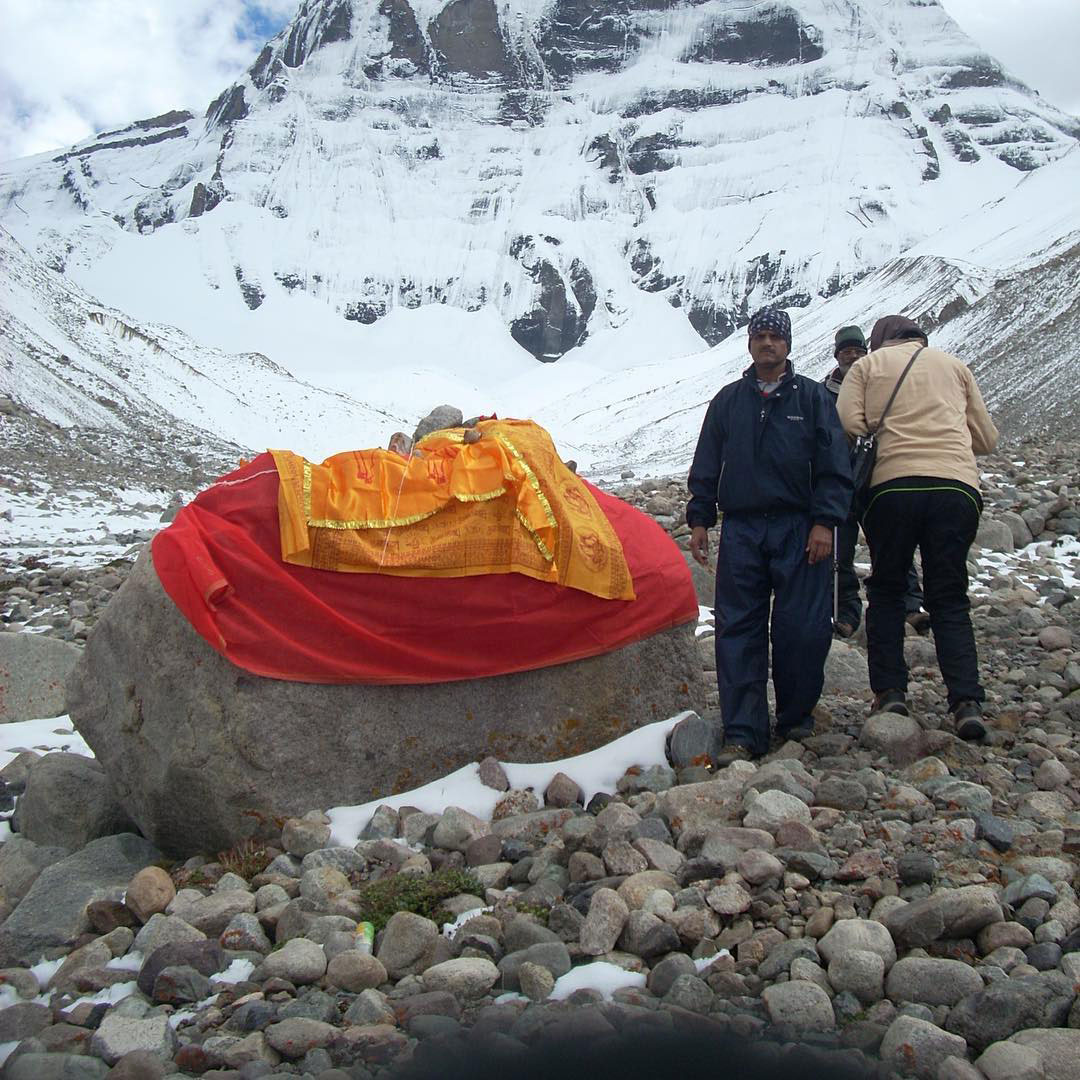 Pilgrims performing rituals at a sacred rock wrapped in prayer cloths during the Mount Kailash Kora, with snow-covered cliffs in the background