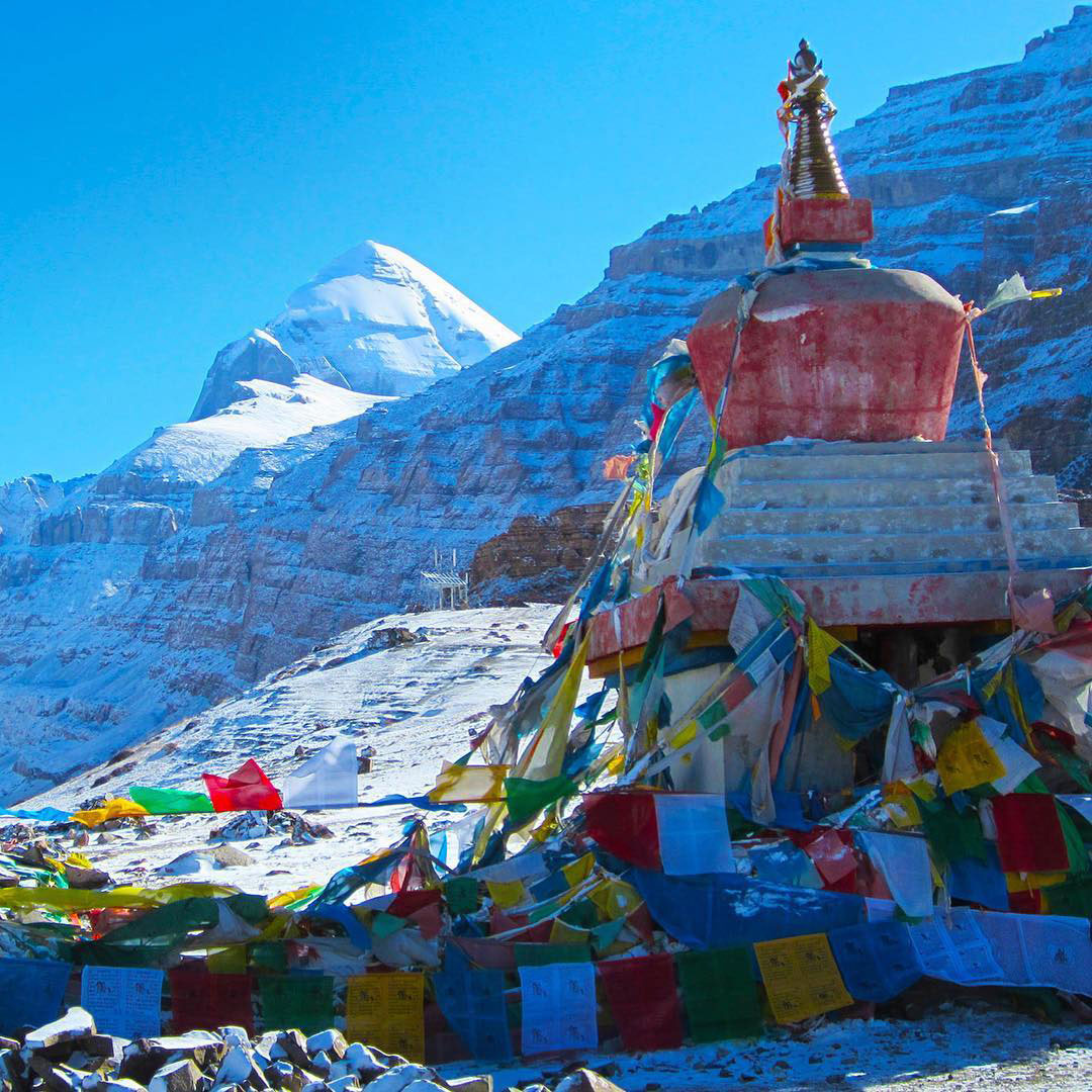 Colorful Tibetan prayer flags surrounding a red stupa with snow-covered Mount Kailash in the background under a bright blue sky