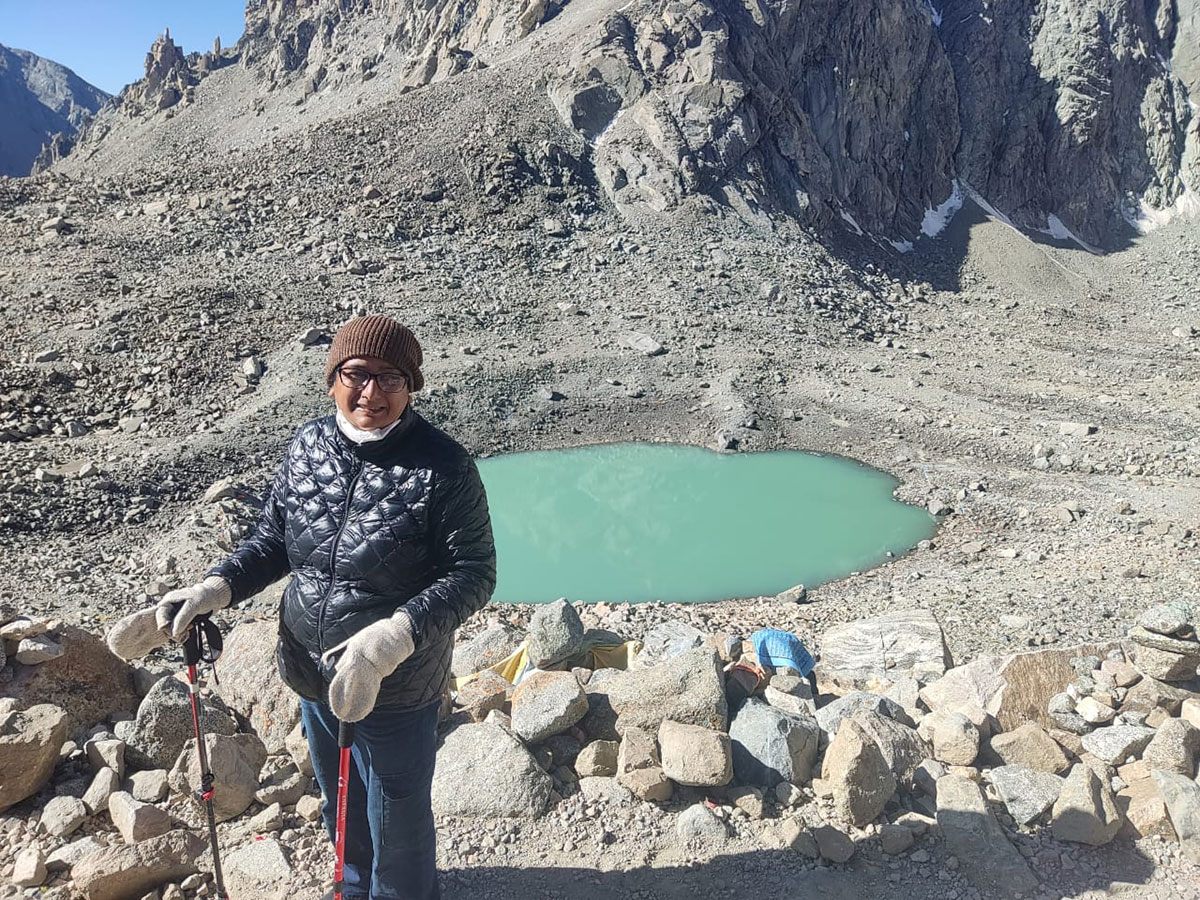 A smiling trekker wearing a winter jacket, gloves, and cap standing near a turquoise glacial lake surrounded by rocky Himalayan mountains.