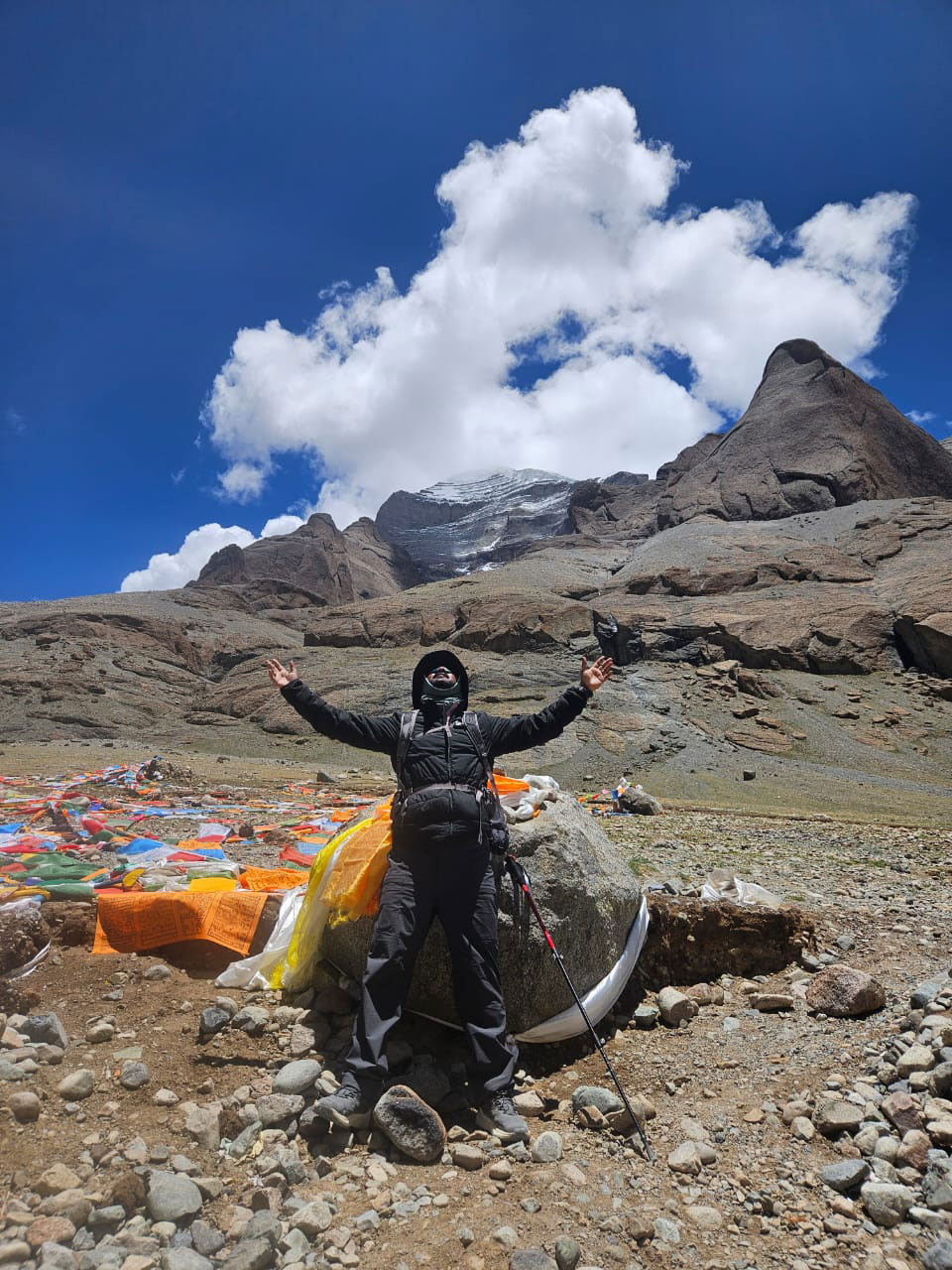 A trekker in black winter gear raising arms in joy near Mount Kailash, surrounded by colorful Tibetan prayer flags under a bright blue sky with white clouds.
